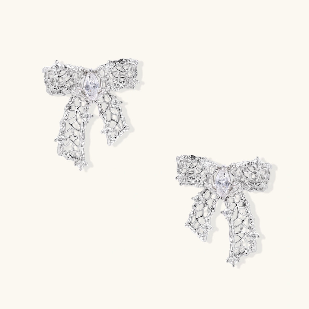 Elegant silver bow-shaped earrings with sparkling zirconia crystals and dangling ribbon details on cream background.