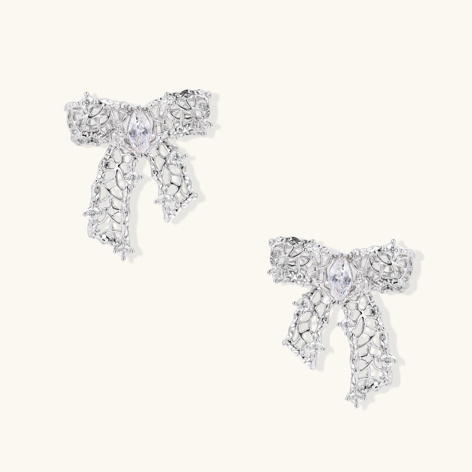 Elegant silver bow-shaped earrings with sparkling zirconia crystals and dangling ribbon details on cream background.